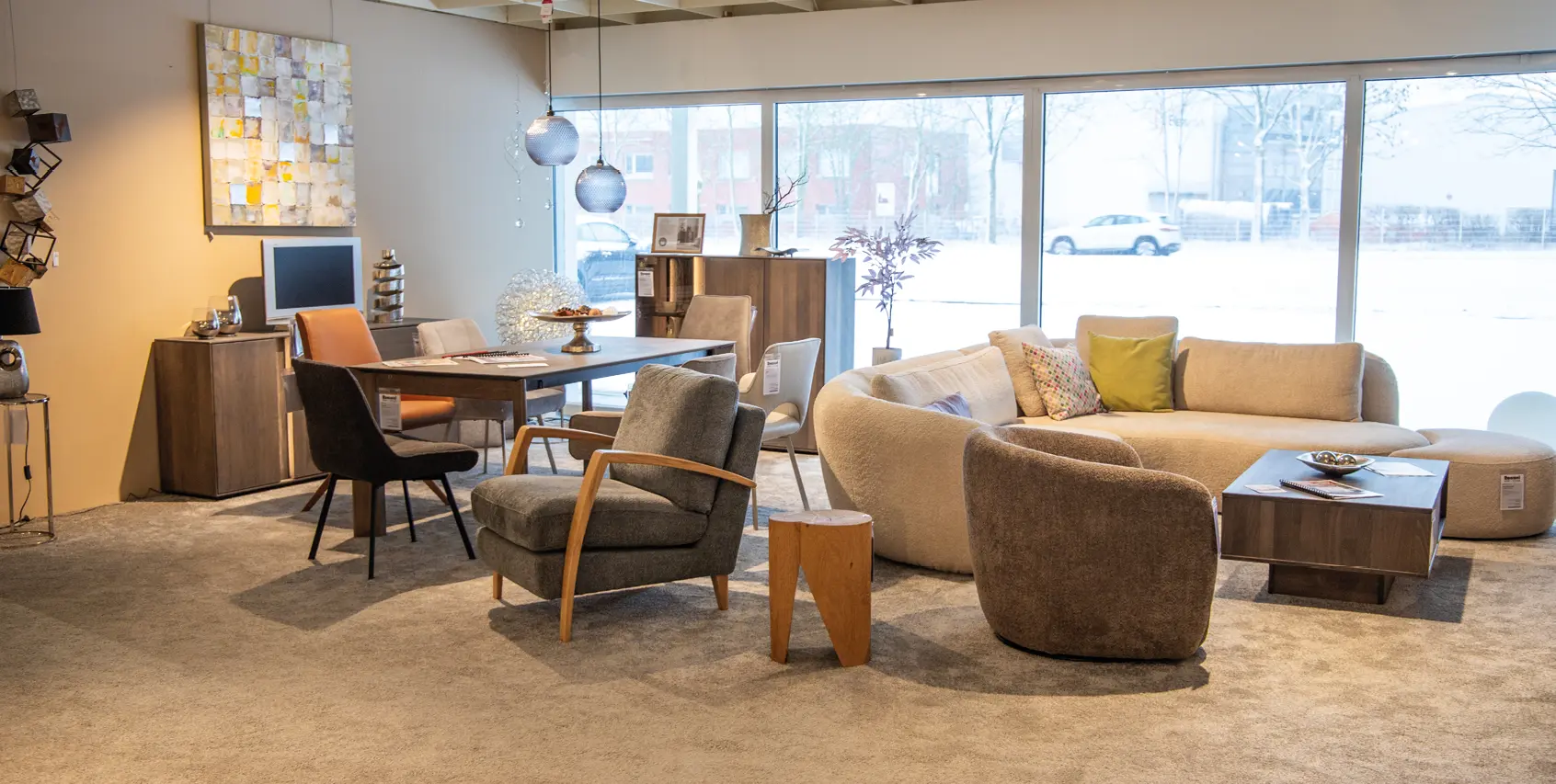 Ausstellung mit hellem Sofa, Sesseln und kleinem Tisch rechts, links Essbereich mit Holztafel und Stühlen, große Fensterfront im Hintergrund und Wanddeko.